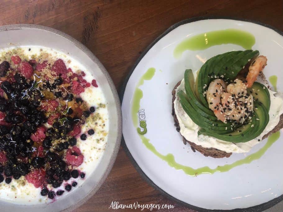 white plate with avocado toast with shrimp and a bowl with yogurt and fruit on wooden table, at In's Myslym Shyri in Tirana