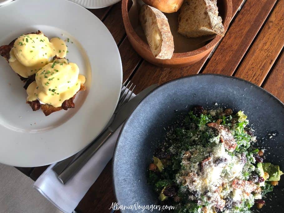eggs benedict on a white plate, kale salad in blue pottery bowl and wooden bowl of bread on wooden table at Lift Steakhouse & Rooftop Bar