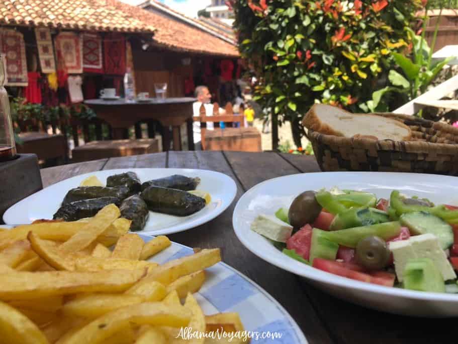 wooden table with plates of fries, salad and japrak and basket of bread with Kruja market in background