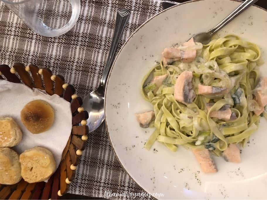 round plate with pasta and chunks of salmon and basket of buns on brown and white place pat in Pasta te Zenga